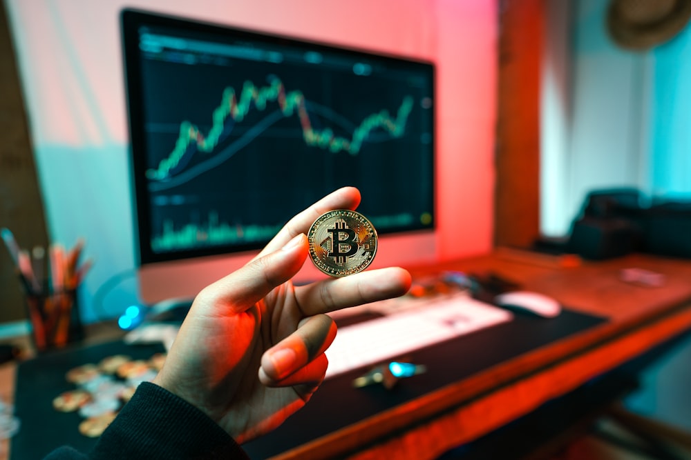 A person holding a Bitcoin coin with trading graphs on a monitor in the background.